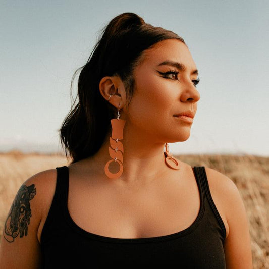 Woman wearing abstract formline art in terracotta color. Top is copper shield, followed by two s shaped formline shapes, and the bottom a circle. earrings from copper canoe woman