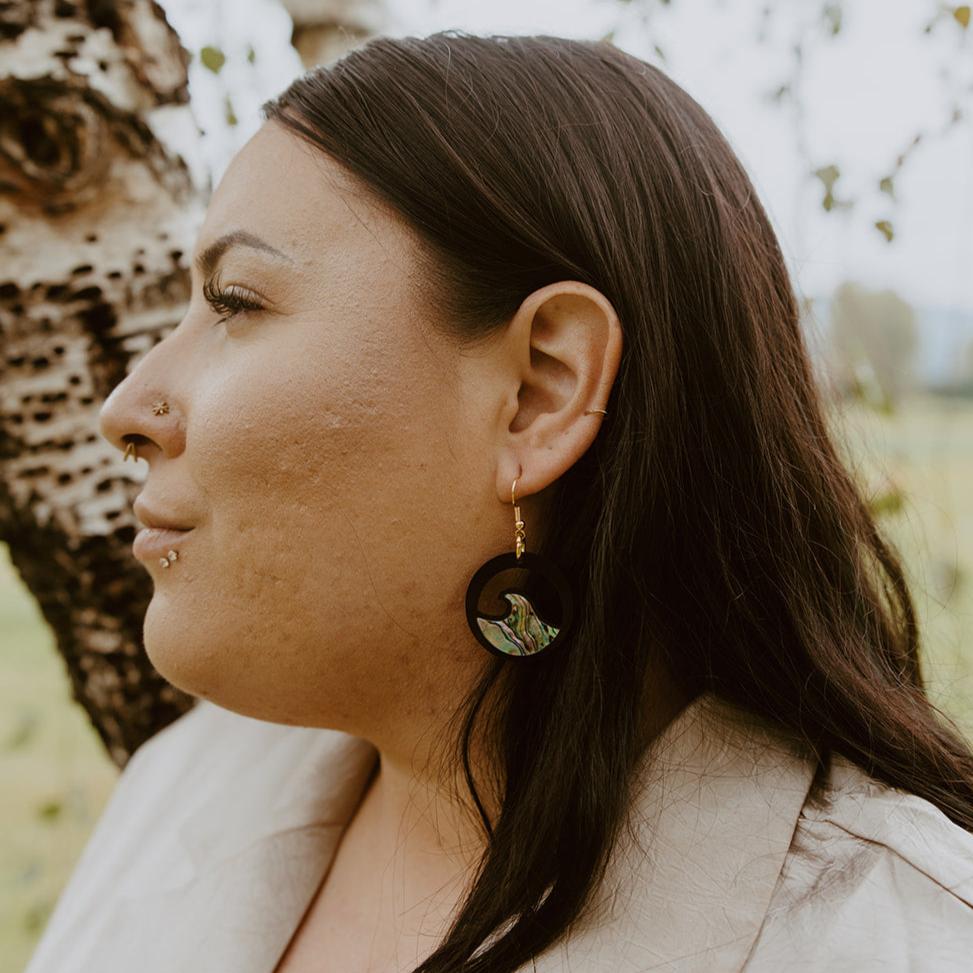 woman wearing black earring with wave cut out and abalone inlay from copper canoe woman