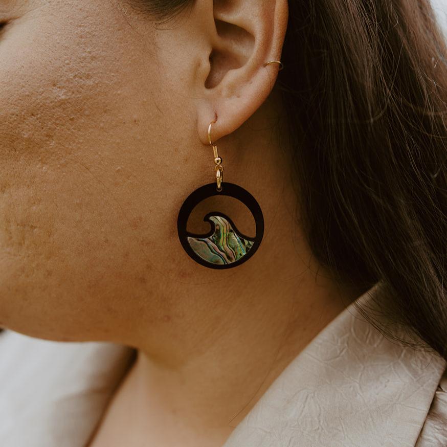 close up of woman wearing black acrylic earring with wave cut out and abalone inlay from copper canoe woman