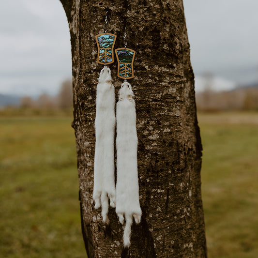 Copper shield earrings with abalone inlay and ermine pelts hang from a tree in a field. Design by Copper Canoe Woman.