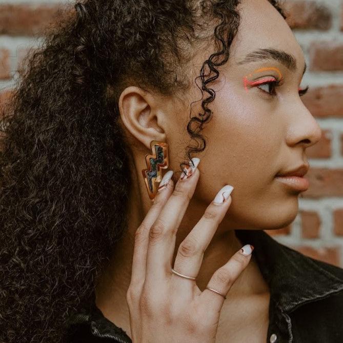 Model wearing the lightning bolt earring from Copper Canoe Woman in gold mirror acrylic with abalone inlay.