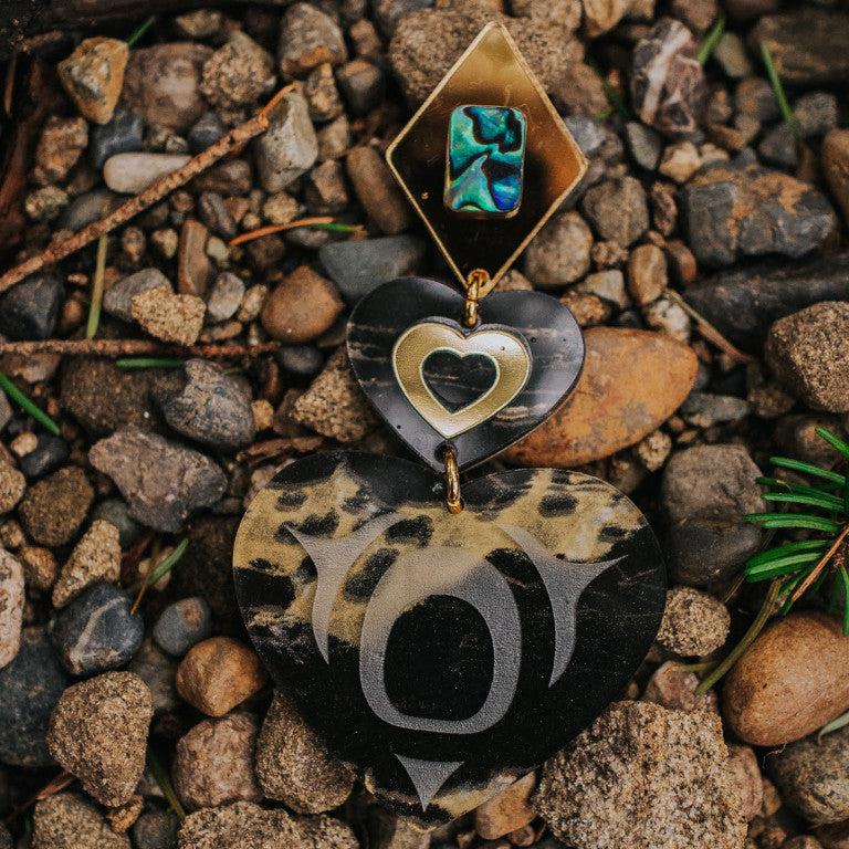 A single monochrome majesty earring in obsidian gold acrylic from Copper Canoe Woman rests on a bed of rocks.