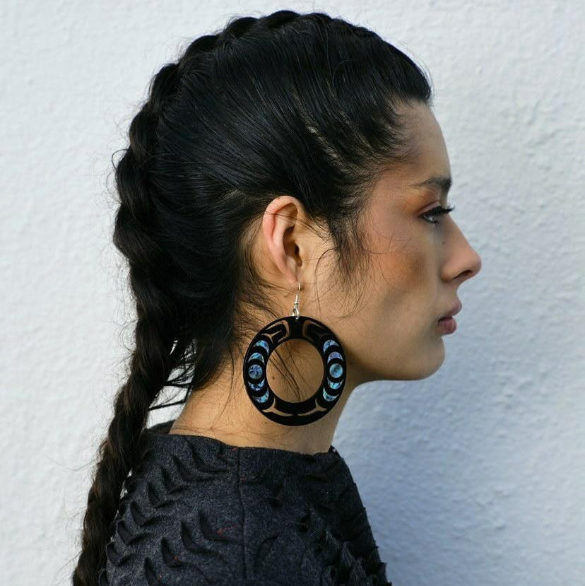 Model stands in profile wearing the Nala Hoop earring from Copper Canoe Woman in black acrylic with abalone inlay.