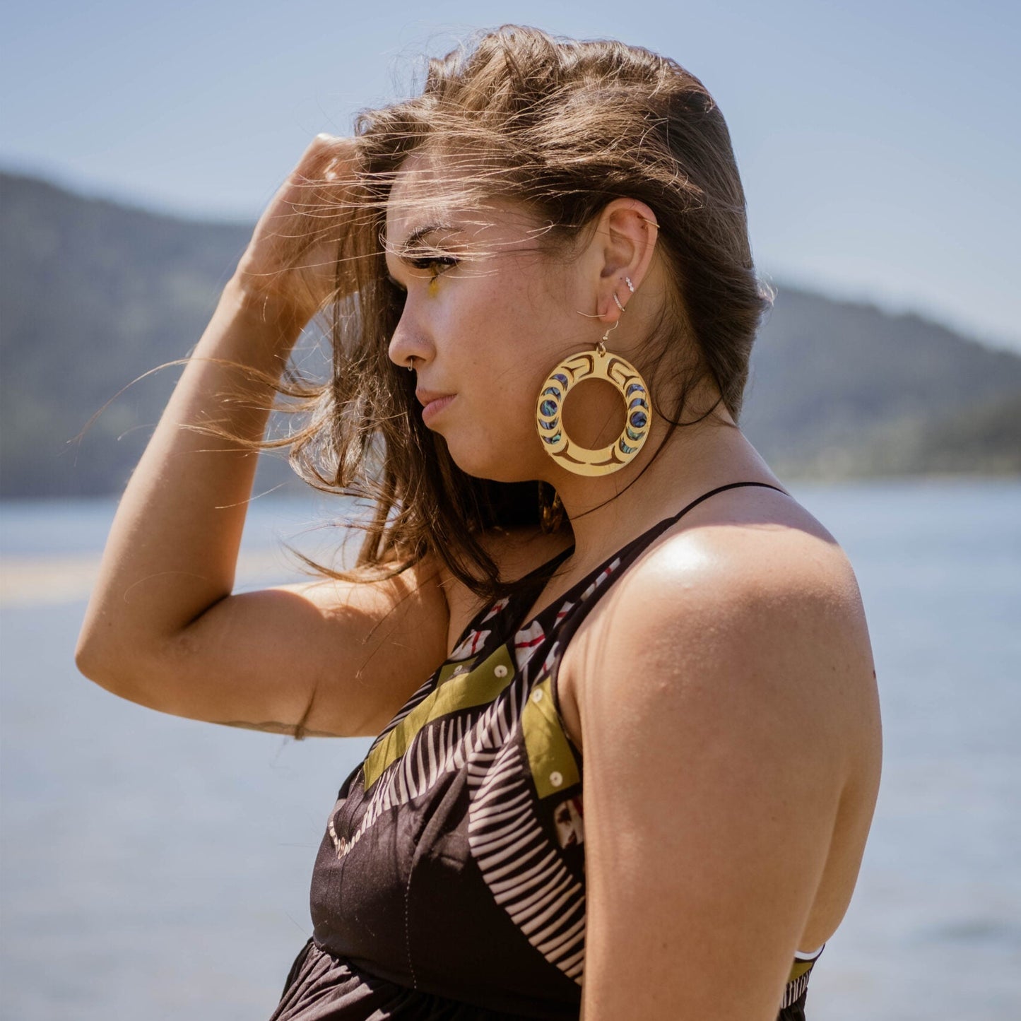 Model standing by water wears the Nala Hoop earring from Copper Canoe Woman in gold mirror acrylic.