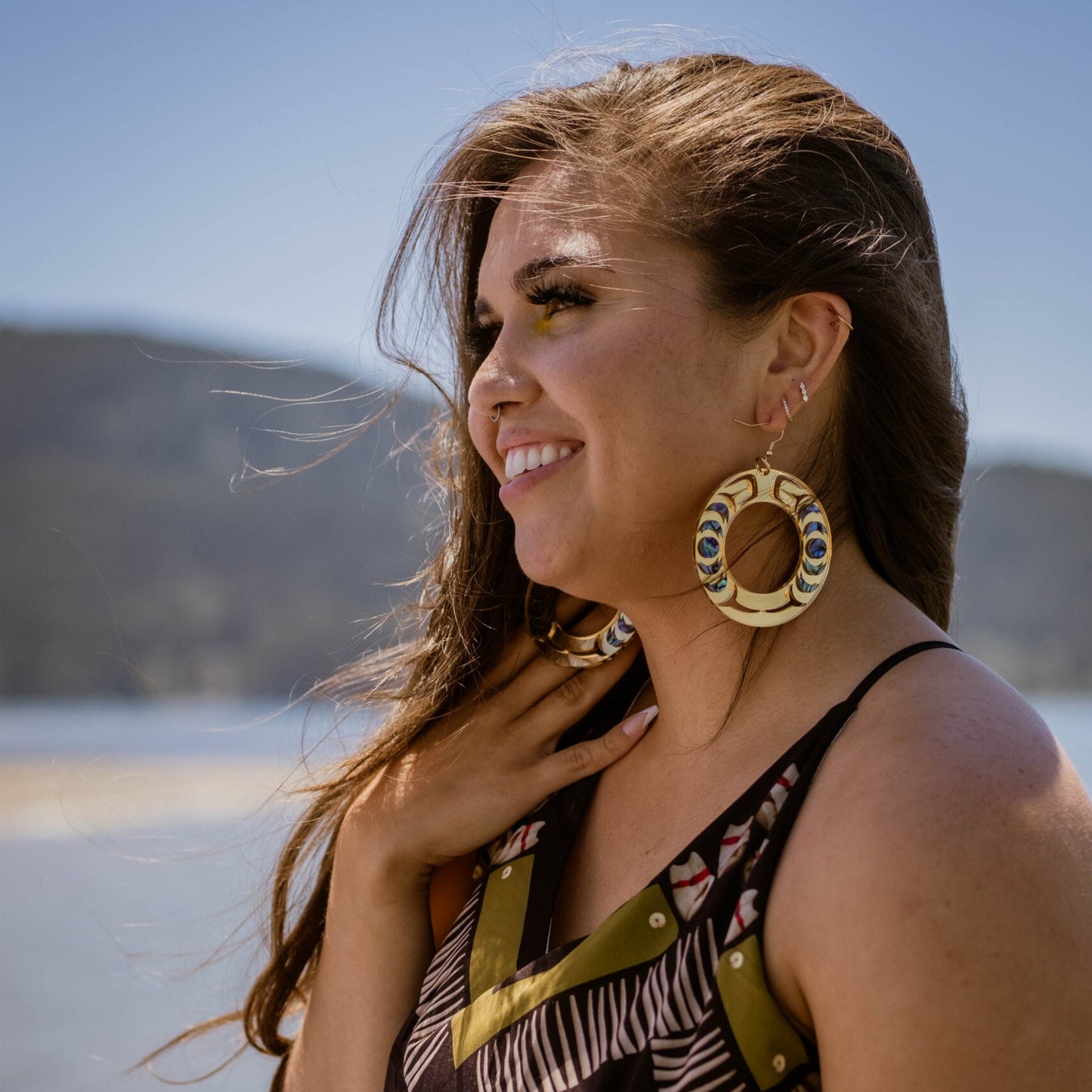 Model standing by water wears Nala Hoop earrings in gold mirror acrylic from Copper Canoe Woman.