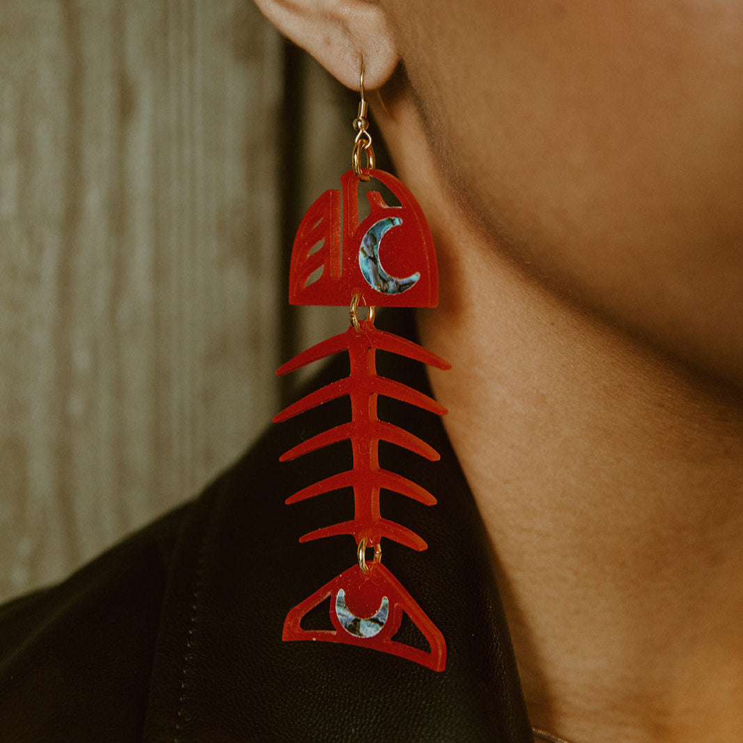 A closeup of a model wearing the Medium Salmon Ghost earrings from Copper Canoe Woman in a red acrylic.