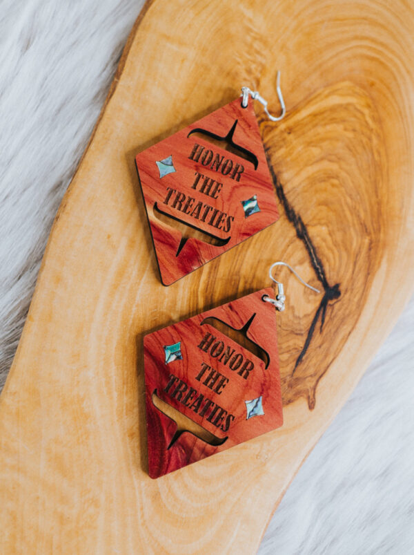 A pair of diamond shaped cedar earring engraved with phrase "Honor the Treaties" rests on a wooden plank.