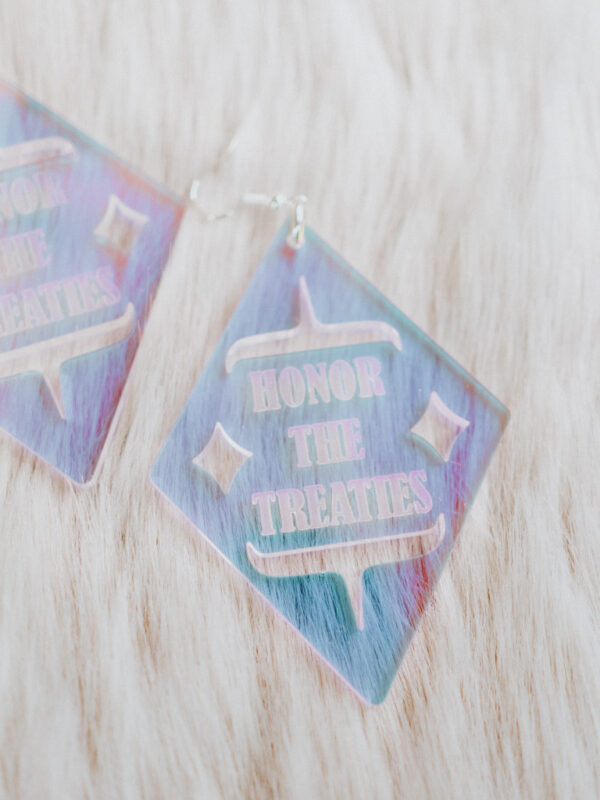 Diamond shaped earrings in iridescent acrylic with cutwork shapes and "Honor the Treaties" cutout letters lay on a white fur.